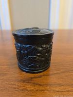 Front view of vintage black round carved resin trinket box showing intricate animal and bird carvings on the side and the lid with a raised design.