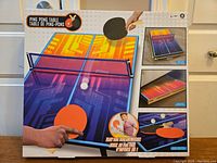 Box showing the Ping Pong Table set with bright colored foldable table, net, paddles, and balls.