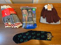 Set of various items including 6-pack men's briefs, brown gloves with knit hat, and black socks with green dollar sign pattern on wooden surface