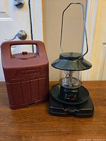 Photo showing green propane lantern next to the brown Coleman carrying case on a wooden surface.