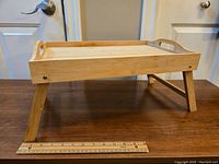 Front side view of foldable bamboo lap tray with legs extended and wooden ruler placed in front for scale, showing smooth tray surface and handles.