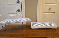 Four white plastic folding shelves with metal legs, stacked and one unfolded showing structure and ventilation holes.