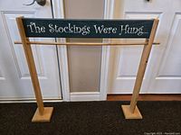 Front view of handmade wooden stocking holder with a green painted plank that reads 'The Stockings Were Hung...' in white whimsical script, supported by two natural wood legs and base.