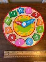 Top down view of Melissa & Doug wooden shape sorting clock showing colored shaped blocks numbered 1 to 12 on outer edge, and blue hour and minute hands in center with minute markings.