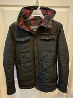 Front view of men's Eddie Bauer black quilted jacket on hanger showing two snap-button chest pockets and attached hood with red and black plaid lining.