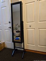 Front view of the black finished wood jewelry armoire standing indoors near door and closet with full-length mirror on door front and two-legged base.