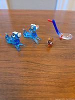 Four Murano glass miniature animal figurines displayed on a wooden surface: two blue dog-like animals, one small brown insect-like figurine, and one long-necked swan.