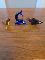 Three miniature Murano glass sea creatures displayed in a row on a wooden surface: amber seahorse, blue dolphin, and brown fish.