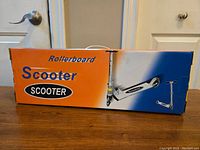 Box of brand new child sized scooter with orange and blue gradient background and 'Scooter' text on box front.