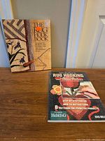 Two books on rug hooking placed on a wood surface, showing titles and cover art with rug hooking images and tools