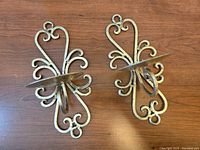 Pair of gold tone metal wall sconces with ornate scrollwork, placed on wooden surface showing their full shape and size.
