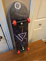 Full view of the sealed skateboard leaning against a wall, showing black deck with graphics, red wheels, and plastic wrapping.