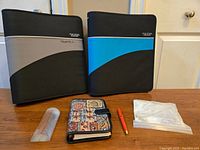 Two Mead Five Star 3-ring zipper binders, a colorful personal planner with black clasp, orange pen, and magnifying glass in plastic case arranged on a flat surface.