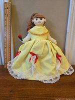 Front view of the doll in yellow gown with lace skirt and red ribbon decorations, holding a flower accessory. Shows overall appearance and size against a ruler.