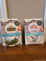 Two cross stitch kits packaged with embroidery hoops, fabric, needle, and threads; one showing a house and the other a rainbow cloud design.