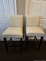 Two cream faux leather bar stools with black wooden legs, standing side by side with plain white door background.