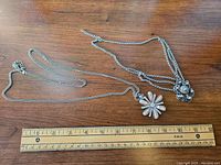 Two necklaces laid flat on a wooden surface with a ruler beneath to show scale, displaying two distinct flower pendants on silver-tone chains.