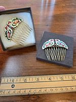Two vintage enamel hair combs shown side by side on a wooden surface with measuring rulers below to show size. One comb has butterfly shaped enamel top and the other fan shaped.