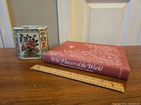 Photo showing the hexagonal floral print tin next to the Wild Flowers of the World book, with a wooden ruler for scale.