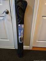 Windshield cover in black carrying bag, leaning against a wall near a door, showing product label with vehicle graphic and text.