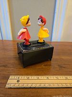 Photo showing the front side of the vintage 1970s plastic kissing couple music box on a wooden surface with a ruler for scale.