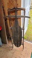 Photo showing front view of antique sled with wooden seat and metal frame, showing weathered condition.
