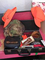 Open suitcase containing hunting safety hats, gloves, leather belt, compasses, pocket cutting tools in leather cases, and a Lexus branded first aid kit.