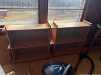 Two pressed wood bookcases placed against a window. Both have two shelves with raised side edges on the top shelf and vertical panel back detail. Medium brown color.