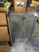 Front view of gray metal cabinet with one drawer and one door showing metal knob and wear marks.