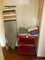 Photo showing full view of vintage red metal rolling cart with three shelves, ironing board standing next to it, and folded towels stacked on the top shelf.