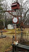 View showing the birdhouse on top of a wooden post with a large round white-faced thermometer below it, a weathered wooden bench below the post with two garden figurines on it, and metal garden chimes hanging from the post next to the birdhouse.