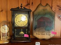 Three items displayed on a wooden shelf: Centurion clock, Elgin anniversary clock under glass dome, Stone Creek Bridge plaque.