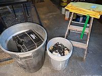 Photo of large black trash bin, smaller gray bin with cords, a wooden stepladder with tarp tied on top, and black crate inside the trash bin.