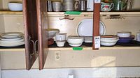 Kitchen cabinet shelf displaying Corelle Spring Blossom Green patterned dinnerware including plates, bowls, and platter, showing quantity and pattern detail.