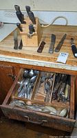 Wooden butcher block with multiple kitchen knives with wooden handles on countertop, drawer open showing assorted spoons, forks, and knives with metal and wooden handles, and serving utensils.