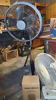 View of the lot showing a large black floor fan, a smaller white tabletop fan, and a boxed TruTech space heater placed on the floor in a basement setting.