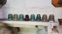 Shelf with eight glass and ceramic insulators in a row showing color and shape variations.
