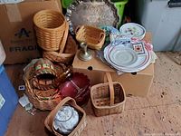 Photo showing multiple Longaberger baskets in different shapes and sizes, some with handles. Also visible are a decorative ceramic floral bowl and other small baskets in front of a cardboard box.