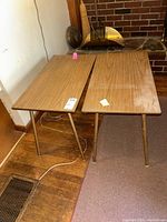 Two vintage Mid-Century Modern style side tables with wood grain tops and metal legs placed adjacent to a brick fireplace and wooden floor, showing overall view and condition.