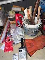 Painting supplies including paint brushes, stir sticks, orange gloves, putty knives, paint thinner bottle, and paint bucket with residue