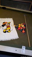 Photo of a billiard table with full set of pool balls in a wooden rack beside a wooden pool cue stick and white towel with Mickey Mouse golfing print
