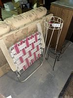 Photo shows the folding lawn chair with red and white woven plastic seat straps and beige fabric backrest standing next to two black metal plant stands, one tall and one short.