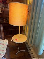 Front view of vintage table lamp turned on, showing beige fabric shade and white metal round table surface with tripod base.