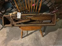 Wooden toolbox on a wooden chair filled with a claw hammer, hand saw, chisels or screwdrivers, wooden plane, and metal file.