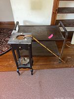 Image showing the metal two-shelf rolling TV stand with caster wheels and the black ashtray stand with ashtray on lower shelf; ashtray stand missing top piece; bamboo cane leaning on the ashtray stand