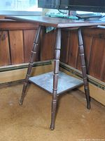 Full view of square wooden accent table with four turned spindle legs and a lower shelf against a wooden-paneled wall. Table surface and legs show scuffs and scratches.