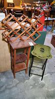 Wide view showing all three items together: wooden plant stand with two small shelves, folding wooden wine rack on top, and vintage folding tray table with decorative top.