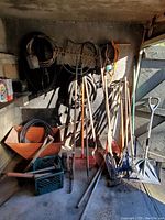 Full view of back patio corner with metal utility cart and multiple yard tools hung on wall and resting against cart. Several coiled hoses seen hung above and around cart. Tools show weathered and used condition.