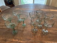 Top-down view of 4 green margarita glasses with thick stems and 6 clear handmade heavy glasses arranged on wooden table.