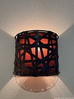 Front view of a single amber and metal wall sconce illuminated on the wall showing the crisscross metal design and warm orange light.